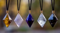 Five colorful crystal pendants hanging on black cords display amber, pink, blue, clear and dark gemstone colors against soft blurred background.