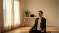 Young Asian woman in business attire practicing mindfulness meditation in modern minimalist home office with natural sunlight streaming through window.