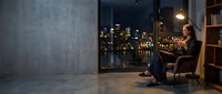 Young professional woman relaxing in modern apartment chair reading book with city skyline view through large windows at night.