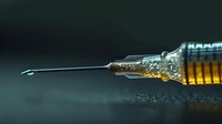 Medical syringe with golden liquid medicine and needle in dramatic lighting against dark background, close-up macro photography showing transparent glass detail.