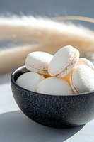 White macarons with delicate filling arranged in textured black ceramic bowl against light background, soft natural lighting creates gentle shadows.