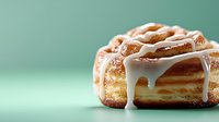 Fresh baked cinnamon roll with sweet white glaze dripping down sides against mint green background, close-up view showing soft layered dough texture and swirls.