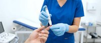 Healthcare professional in blue scrubs performing blood glucose test on patient finger using lancet device in modern medical clinic setting.