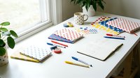 Flat lay workspace with patterned notebooks, blank notepad, markers, and office supplies arranged on white desk near window with potted plants.