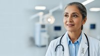 Professional female doctor with stethoscope smiling confidently in modern hospital corridor. Healthcare worker portrait for medical marketing materials.