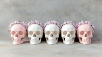 Pink and white decorative skulls adorned with delicate floral crowns arranged in row against neutral background, creating modern gothic aesthetic for seasonal decor.