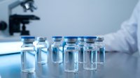 Glass vials with blue caps arranged on a lab table, microscope and scientist in white coat blurred in background.