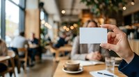 Young adult male of mixed ethnicity holding blank white business card in modern cafe setting with blurred background, suitable for corporate branding and networking concepts.