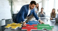 Professional businessman in blue suit assembling colorful puzzle pieces labeled with business departments in modern conference room with diverse team.