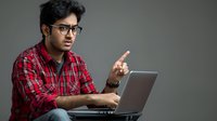 Young South Asian man in red plaid shirt with glasses pointing while working on laptop against gray background, expressing discovery or instruction.