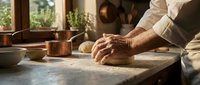 Mature male baker kneading dough on wooden surface in rustic kitchen with natural light and copper cookware visible in background.