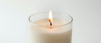 Lit white candle in a clear glass jar with a glowing flame, soft warm light on a neutral background for spa and wellness use.