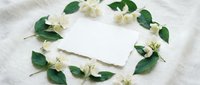 Blank white card surrounded by fresh jasmine flowers and green leaves arranged in a circle on linen fabric, ready for wedding invitations, greeting cards, or stationery design projects.