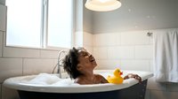Young African American woman relaxing in bathtub with rubber duck toy, enjoying peaceful bath time in modern bathroom with natural light and towels.