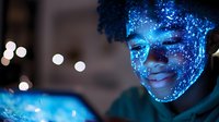 Young African American person illuminated by blue digital projection mapping on face showing bright smile, looking at glowing device screen in dark room.