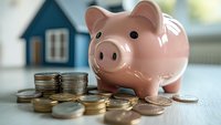 Pink ceramic piggy bank with stacked coins and small house model in background, representing savings, mortgage and financial planning for home ownership.