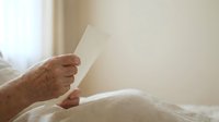 Senior adult male with aged skin holding blank white envelope while sitting on bed in bright bedroom setting.