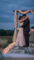 Romantic newlywed couple embracing at twilight wedding ceremony with glowing string lights on rustic wooden arch and deck for outdoor celebration.