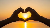 Hands forming heart shape silhouette against golden sunset over calm water creating romantic peaceful moment for love wellness meditation concepts.