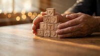 Hands protecting wooden blocks with heart icons stacked in pyramid shape on table with warm bokeh lights background for love care protection concept.