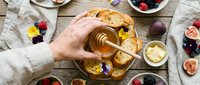Hand drizzling honey onto toasted bread with butter, surrounded by fresh berries and figs on rustic wooden table for food styling and culinary content.