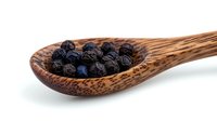 Dried black peppercorns in rustic wooden spoon on white background, macro food photography showing texture and detail of aromatic spice ingredient.