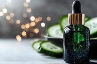Green glass dropper bottle with sparkly serum against fresh cucumber slices and soft bokeh lights, showcasing natural skincare and beauty product photography.
