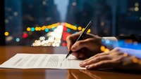 Professional hand signing important contract document with pen at wooden desk during evening hours with colorful city lights bokeh background.