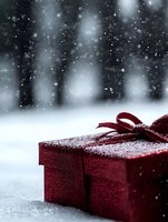 Red gift box with bow in snow during snowfall, creating magical holiday atmosphere. Festive present against blurred winter forest background.