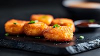 Golden crispy potato croquettes garnished with fresh herbs on black stone surface, selective focus with blurred sauce bowl in background. Appetizing comfort food.