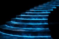 Glowing blue neon spiral staircase steps illuminated in darkness with ethereal light effect creating futuristic architectural detail against black background.