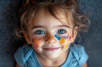 Smiling young child with bright blue eyes wearing face paint with colorful hearts and musical notes design on cheeks, dressed in light blue shirt against dark background.