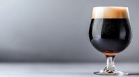 Dark craft beer in snifter glass with creamy foam head and condensation drops against gray background, dramatic side lighting creates appetizing mood.