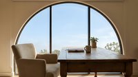 Modern minimalist workspace with wooden desk, beige armchair, and potted plant framed by large arched window overlooking natural landscape, ideal for interior design and office concepts.