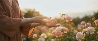 Woman in beige cardigan with open hands in peaceful gesture among blooming roses during golden hour sunset creating serene mindfulness moment for wellness content.