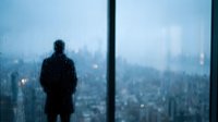 Silhouette of businessman standing by office window overlooking city skyline on rainy evening with water droplets and urban lights creating moody atmosphere.