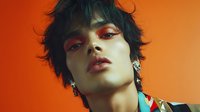 Young Asian man with tousled black hair and bold red eyeshadow makeup against vibrant orange background for fashion and beauty campaigns.