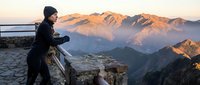 Photographer in black jacket and beanie capturing mountain landscape at golden hour from rocky overlook, scenic alpine peaks and valley below at sunrise or sunset.
