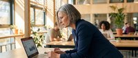 Senior woman with gray hair, Caucasian ethnicity, wearing dark blazer, focused on laptop screen in modern office environment with colleagues working in background.