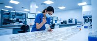 Female laboratory technician of Asian descent wearing blue scrubs and protective mask examining samples in modern medical research facility with advanced equipment and sterile environment.