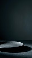 Empty black plate on dark textured surface with dramatic side lighting, creating strong shadows. Suitable for food styling, restaurant menus, culinary presentations, and minimalist design projects.