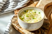 Creamy potato leek soup garnished with fresh chives and black pepper in rustic ceramic bowl, served on wooden board with striped linen napkin. Natural lighting.