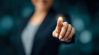 Businesswoman in formal attire pointing finger at virtual interface against blurred blue background, demonstrating digital technology interaction.