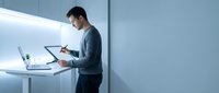 Young Asian businessman writing notes while working on laptop at modern standing desk in minimalist office with LED lighting for productivity concepts.
