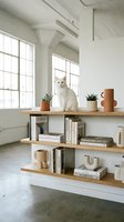 White cat sitting on wooden shelf unit with books, plants and decorative objects in minimalist interior with large windows and industrial ceiling.