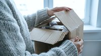 Woman in gray sweater holding open empty cardboard box near bright window, unboxing or unpacking delivery package indoors.