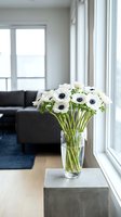 White anemone flowers in glass vase on concrete table by window in modern living room with natural light and contemporary furniture for home decor.