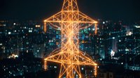 Illuminated electrical transmission tower glowing with orange light against dark city skyline at night. Urban infrastructure and power grid concept for energy industry.