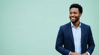 Young African American male professional wearing navy blazer and white shirt, smiling confidently against mint green background. Suitable for corporate, business, and recruitment marketing materials.