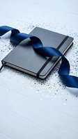 Gray notebook with navy blue satin ribbon and colorful confetti on white wooden surface for celebration planning and gift wrapping concepts.